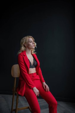Young adult beauty woman in formal evening suit of red color with lace black bra underwear standing by thoughtful. Stylish blonde curly hair model fashionista posing at studio in fashion pantsuit