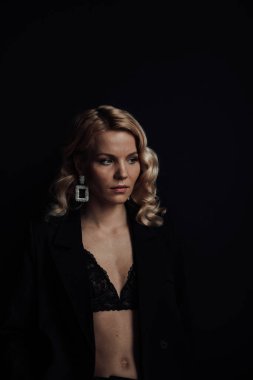 Young adult beauty woman in formal evening suit of black color with lace bra at thoughtful. Stylish blonde curly hair sensual model fashionista posing at studio in fashion pantsuit