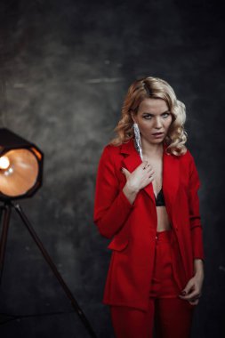 Young adult beauty woman in formal evening suit of red color with lace black bra underwear standing by thoughtful. Stylish blonde curly hair model fashionista posing at studio in fashion pantsuit