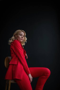 Young adult beauty woman in formal evening suit of red color with lace black bra underwear standing by thoughtful. Stylish blonde curly hair model fashionista posing at studio in fashion pantsuit