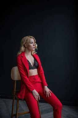 Young adult beauty woman in formal evening suit of red color with lace black bra underwear standing by thoughtful. Stylish blonde curly hair model fashionista posing at studio in fashion pantsuit