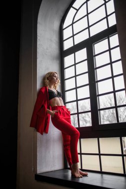 Young adult beauty woman in formal evening suit of red color with lace black bra underwear standing by thoughtful. Stylish blonde curly hair model fashionista posing at studio in fashion pantsuit