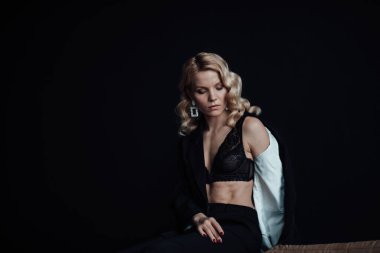 Young adult beauty woman in formal evening suit of black color with lace bra at thoughtful. Stylish blonde curly hair sensual model fashionista posing at studio in fashion pantsuit