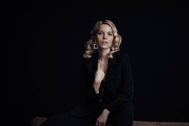 Young adult beauty woman in formal evening suit of black color with lace bra at thoughtful. Stylish blonde curly hair sensual model fashionista posing at studio in fashion pantsuit