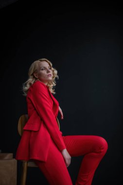 Young adult beauty woman in formal evening suit of red color with lace black bra underwear standing by thoughtful. Stylish blonde curly hair model fashionista posing at studio in fashion pantsuit