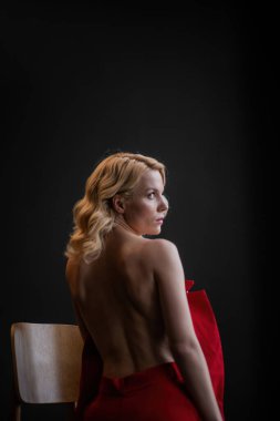 Adult beauty woman bare back in formal evening red trousers sitting pose without bra. Stylish blonde curly hair sensual nude model fashionista posing at studio in fashion pantsuit out of blazer
