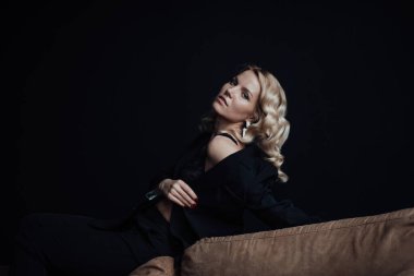 Young adult beauty woman in formal evening suit of black color with lace bra at thoughtful. Stylish blonde curly hair sensual model fashionista posing at studio in fashion pantsuit