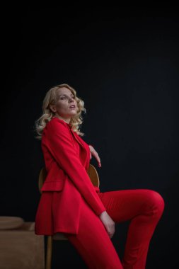 Young adult beauty woman in formal evening suit of red color with lace black bra underwear standing by thoughtful. Stylish blonde curly hair model fashionista posing at studio in fashion pantsuit