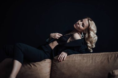 Young adult beauty woman in formal evening suit of black color with lace bra at thoughtful. Stylish blonde curly hair sensual model fashionista posing at studio in fashion pantsuit