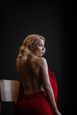 Adult beauty woman bare back in formal evening red trousers sitting pose without bra. Stylish blonde curly hair sensual nude model fashionista posing at studio in fashion pantsuit out of blazer