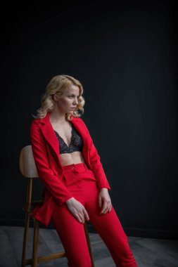 Young adult beauty woman in formal evening suit of red color with lace black bra underwear standing by thoughtful. Stylish blonde curly hair model fashionista posing at studio in fashion pantsuit