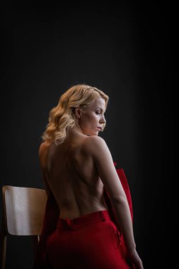 Adult beauty woman bare back in formal evening red trousers sitting pose without bra. Stylish blonde curly hair sensual nude model fashionista posing at studio in fashion pantsuit out of blazer