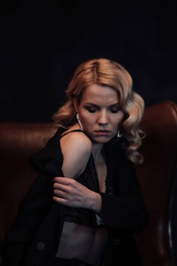 Young adult beauty woman in formal evening suit of black color with lace bra at thoughtful. Stylish blonde curly hair sensual model fashionista posing at studio in fashion pantsuit