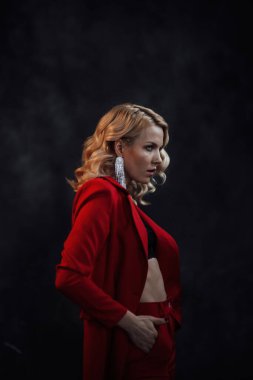 Young adult beauty woman in formal evening suit of red color with lace black bra underwear standing by thoughtful. Stylish blonde curly hair model fashionista posing at studio in fashion pantsuit