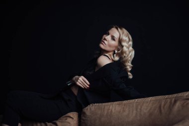 Young adult beauty woman in formal evening suit of black color with lace bra at thoughtful. Stylish blonde curly hair sensual model fashionista posing at studio in fashion pantsuit