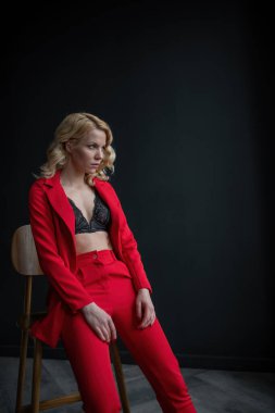 Young adult beauty woman in formal evening suit of red color with lace black bra underwear standing by thoughtful. Stylish blonde curly hair model fashionista posing at studio in fashion pantsuit