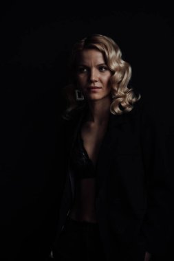 Young adult beauty woman in formal evening suit of black color with lace bra at thoughtful. Stylish blonde curly hair sensual model fashionista posing at studio in fashion pantsuit