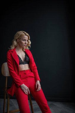 Young adult beauty woman in formal evening suit of red color with lace black bra underwear standing by thoughtful. Stylish blonde curly hair model fashionista posing at studio in fashion pantsuit