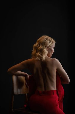 Adult beauty woman bare back in formal evening red trousers sitting pose without bra. Stylish blonde curly hair sensual nude model fashionista posing at studio in fashion pantsuit out of blazer