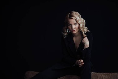 Young adult beauty woman in formal evening suit of black color with lace bra at thoughtful. Stylish blonde curly hair sensual model fashionista posing at studio in fashion pantsuit