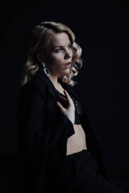 Young adult beauty woman in formal evening suit of black color with lace bra at thoughtful. Stylish blonde curly hair sensual model fashionista posing at studio in fashion pantsuit