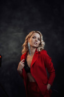 Young adult beauty woman in formal evening suit of red color with lace black bra underwear standing by thoughtful. Stylish blonde curly hair model fashionista posing at studio in fashion pantsuit