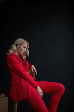 Young adult beauty woman in formal evening suit of red color with lace black bra underwear standing by thoughtful. Stylish blonde curly hair model fashionista posing at studio in fashion pantsuit