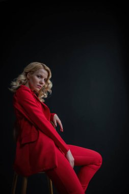 Young adult beauty woman in formal evening suit of red color with lace black bra underwear standing by thoughtful. Stylish blonde curly hair model fashionista posing at studio in fashion pantsuit