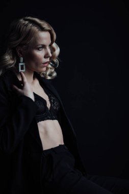 Young adult beauty woman in formal evening suit of black color with lace bra at thoughtful. Stylish blonde curly hair sensual model fashionista posing at studio in fashion pantsuit