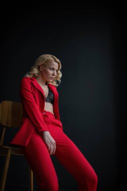 Young adult beauty woman in formal evening suit of red color with lace black bra underwear standing by thoughtful. Stylish blonde curly hair model fashionista posing at studio in fashion pantsuit