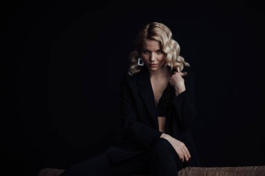 Young adult beauty woman in formal evening suit of black color with lace bra at thoughtful. Stylish blonde curly hair sensual model fashionista posing at studio in fashion pantsuit