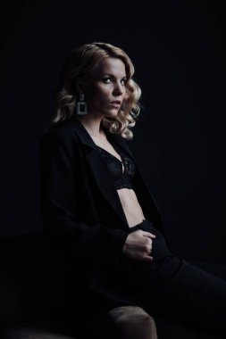 Young adult beauty woman in formal evening suit of black color with lace bra at thoughtful. Stylish blonde curly hair sensual model fashionista posing at studio in fashion pantsuit