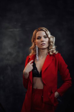 Young adult beauty woman in formal evening suit of red color with lace black bra underwear standing by thoughtful. Stylish blonde curly hair model fashionista posing at studio in fashion pantsuit