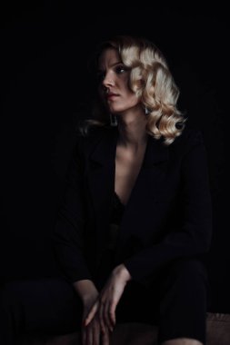 Young adult beauty woman in formal evening suit of black color with lace bra at thoughtful. Stylish blonde curly hair sensual model fashionista posing at studio in fashion pantsuit