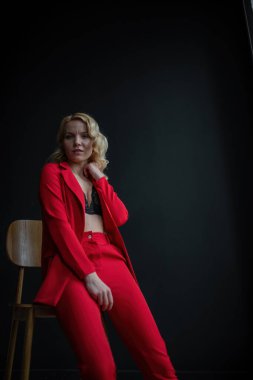 Young adult beauty woman in formal evening suit of red color with lace black bra underwear standing by thoughtful. Stylish blonde curly hair model fashionista posing at studio in fashion pantsuit