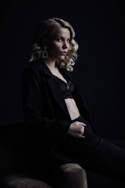Young adult beauty woman in formal evening suit of black color with lace bra at thoughtful. Stylish blonde curly hair sensual model fashionista posing at studio in fashion pantsuit