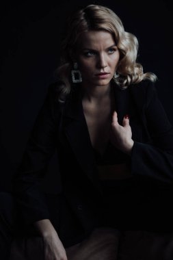Young adult beauty woman in formal evening suit of black color with lace bra at thoughtful. Stylish blonde curly hair sensual model fashionista posing at studio in fashion pantsuit