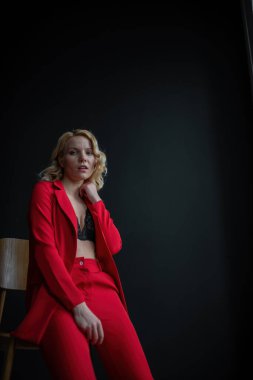 Young adult beauty woman in formal evening suit of red color with lace black bra underwear standing by thoughtful. Stylish blonde curly hair model fashionista posing at studio in fashion pantsuit