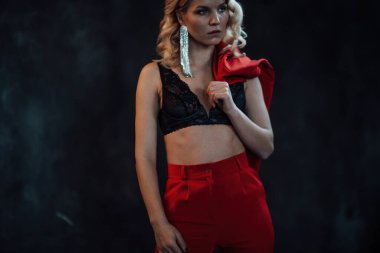 Young adult beauty woman in formal evening suit of red color with lace black bra underwear standing by thoughtful. Stylish blonde curly hair model fashionista posing at studio in fashion pantsuit