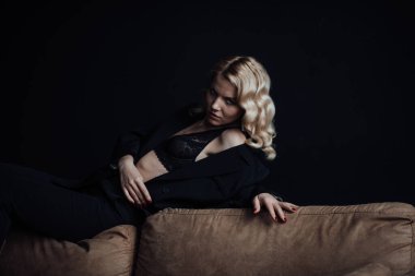 Young adult beauty woman in formal evening suit of black color with lace bra at thoughtful. Stylish blonde curly hair sensual model fashionista posing at studio in fashion pantsuit