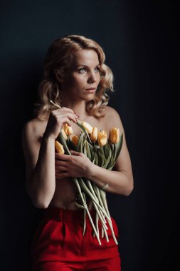 Adult beauty woman half naked in formal evening red trousers without bra hugs bouquet of yellow tulips. Stylish blonde curly hair sensual nude model fashionista posing in studio at spring holidays clipart