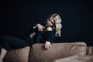 Young adult beauty woman in formal evening suit of black color with lace bra at thoughtful. Stylish blonde curly hair sensual model fashionista posing at studio in fashion pantsuit