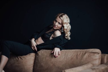 Young adult beauty woman in formal evening suit of black color with lace bra at thoughtful. Stylish blonde curly hair sensual model fashionista posing at studio in fashion pantsuit