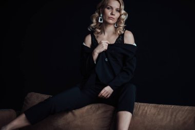 Young adult beauty woman in formal evening suit of black color with lace bra at thoughtful. Stylish blonde curly hair sensual model fashionista posing at studio in fashion pantsuit