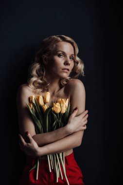 Adult beauty woman half naked in formal evening red trousers without bra hugs bouquet of yellow tulips. Stylish blonde curly hair sensual nude model fashionista posing in studio at spring holidays