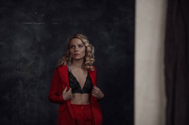 Young adult beauty woman in formal evening suit of red color with lace black bra underwear standing by thoughtful. Stylish blonde curly hair model fashionista posing at studio in fashion pantsuit