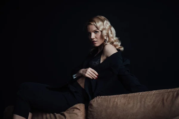 Young adult beauty woman in formal evening suit of black color with lace bra at thoughtful. Stylish blonde curly hair sensual model fashionista posing at studio in fashion pantsuit