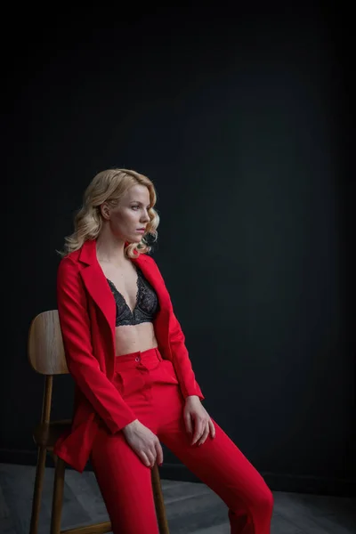 Young adult beauty woman in formal evening suit of red color with lace black bra underwear standing by thoughtful. Stylish blonde curly hair model fashionista posing at studio in fashion pantsuit