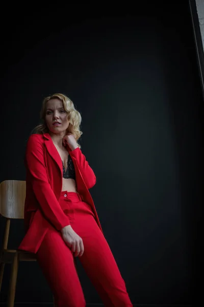 Young adult beauty woman in formal evening suit of red color with lace black bra underwear standing by thoughtful. Stylish blonde curly hair model fashionista posing at studio in fashion pantsuit