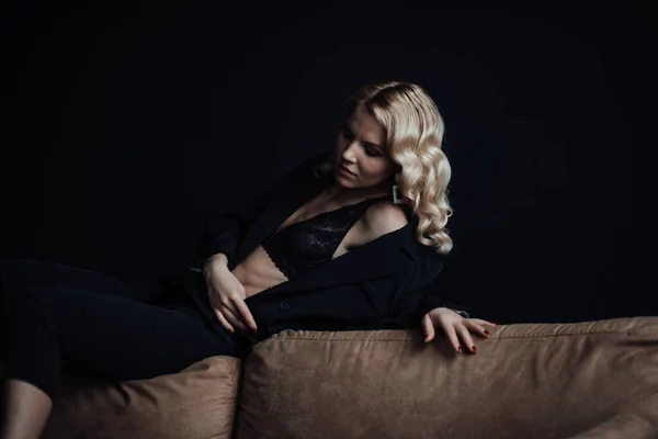 Young adult beauty woman in formal evening suit of black color with lace bra at thoughtful. Stylish blonde curly hair sensual model fashionista posing at studio in fashion pantsuit